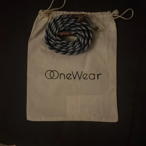 Randigt bälte från OneWear - Säljer ett snyggt randigt bälte från OneWear i blått, vitt och svart. Bältet har en flätad design och metallspänne som ger en cool och modern look. Perfekt för att lyfta din outfit med lite extra stil.