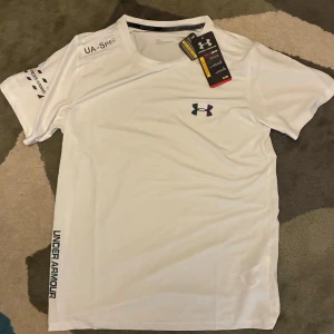 Vit Under Armour HeatGear t-shirt - Snygg vit t-shirt från Under Armour med coolt logotryck i regnbågsfärger på bröstet och längs sidan. Tillverkad i lätt och ventilerande polyester, perfekt för träning. Kortärmad modell med rund hals och tryck på ärmen. THREADBORNE HeatGear-teknologi för extra komfort.