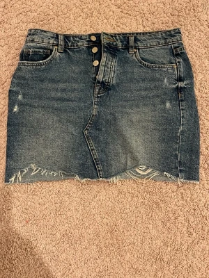 Blå jeanskjol från ONLY med slits - Säljer en blå jeanskjol från ONLY med rå kant och slitningar längst ner. Kjolen har fem fickor, knappstängning framtill och bälteshällor. Klassisk denimlook med snygga detaljer och en liten slits framtill. Perfekt för dig som gillar avslappnad stil.