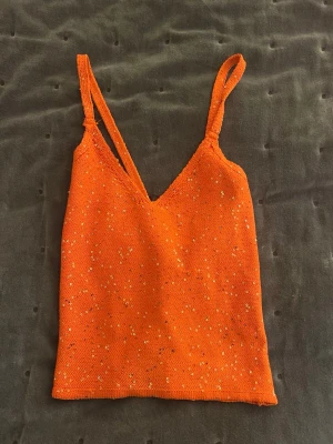 Orange paljettlinnetopp med smala axelband - Säljer en orange linnetopp med smala axelband som korsas i ryggen. Toppen är dekorerad med små paljetter som ger ett glittrigt intryck. Materialet är stickat och toppen har en v-ringad front. Perfekt för sommarens alla tillfällen!