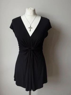 Svart omlottopp med knyt i midjan - Säljer en svart omlottopp med v-ringning och knyt i midjan. Toppen är ärmlös och har en snygg drapering framtill som ger en cool siluett Den är perfekt för att styla med smycken eller lager på lager💕passar xs - s. Har inga defekter 