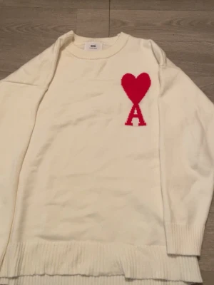 Vit stickad tröja från Ami  - Säljer en vit stickad tröja från Ami med ett stort rött hjärta och bokstaven A på bröstet. Tröjan har rund hals, ribbade muddar och är långärmad. Materialet är mjukt och skönt, perfekt för kyliga dagar. Ikonisk design som sticker ut.