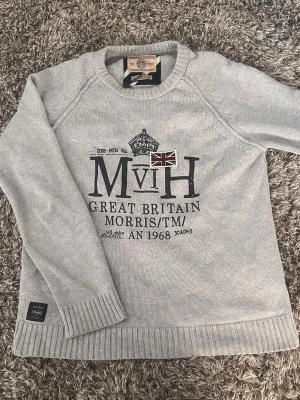 Grå stickad tröja Morris✅🤩 - Snygg grå stickad tröja från Morris Gents med tryck och broderad brittisk flagga på bröstet. Tröjan har rund hals, ribbade muddar och nederkant samt lång ärm. Perfekt för dig som gillar klassisk stil med en twist.🙌🤩