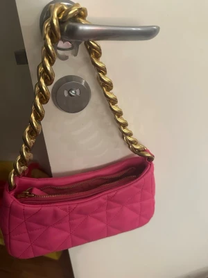 Rosa quiltad axelväska från MNG - Säljer en snygg rosa axelväska från MNG med quiltad design och grov guldfärgad kedja som axelrem. Väskan har dragkedja upptill och är tillverkad i syntetmaterial. Perfekt för dig som vill sticka ut med färg och coola detaljer.