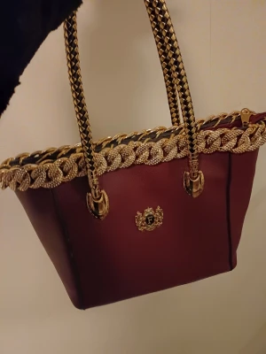 Vinröd handväska med gulddetaljer - Säljer en vinröd handväska med lyxiga guldfärgade kedjehandtag och dekorativt emblem framtill. 