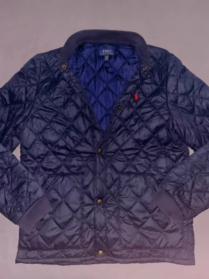 Mörkblå quiltad jacka,Ralph Lauren - Storlek: xs/s.                                                Skick:mycket bra.                                      Beskrivning:Quiltad mörkblå jacka från Polo Ralph Lauren med klassisk krage och tryckknappar framtill. Jackan har ett rött broderat Polo-logo på bröstet och ribbade muddar vid ärmslut. Perfekt för dig som vill ha en stilren och bekväm jacka.