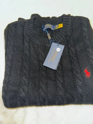 Mörkgrå stickad tröja Polo Ralph Lauren - Snygg mörkgrå stickad tröja från Polo Ralph Lauren med klassiskt kabelstickat mönster och rund hals. Tröjan har lång ärm och den ikoniska röda Polo-loggan broderad på bröstet. Materialmix av ull, akryl och nylon för skön känsla. Använd 1 gång.