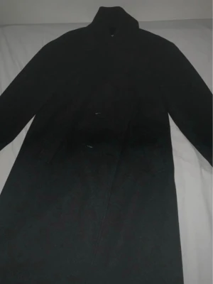 Svart lång kappa med knappar - Säljer en svart lång kappa med dubbelknäppning och klassisk krage. Kappan har en rak och oversized passform med långa ärmar och två sidofickor. Materialet är ullblandning, perfekt för kyliga dagar.