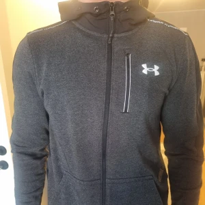 Mörkgrå hoodie från Under Armour - Mörkgrå hoodiejacka från Under Armour med dragkedja och vit logga på bröstet. Jackan har huva med snörning och två sidofickor. Se gärna mina andra annonser och skapa bundles. Inga byten. 