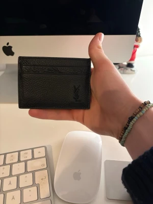Svart Saint Laurent plånbok i skinn - Säljer en stilren svart plånbok från Saint Laurent. Plånboken har 4, kortfack och den ikoniska YSL-loggan framtill. Perfekt storlek för att ha med sig överallt och kommer med originalask och äkthetskort.