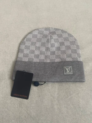 Grå rutig Louis Vuitton mössa - Snygg grå mössa från Louis Vuitton med klassiskt rutigt mönster och ett broderat LV-emblem framtill. Mössan är stickad och har en bred uppvikt kant som ger extra stilpoäng. Perfekt accessoar för att lyfta din look under kyliga dagar.