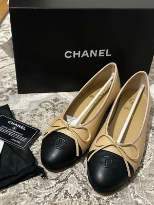 Beige och svarta ballerinaskor från Chanel - Lyxiga ballerinaskor från Chanel i beige och svart skinn med rund tå och dekorativ rosett framtill. Ikoniska CC-loggan är broderad på den svarta tån. Skorna är tillverkade i Italien och levereras med originalkartong och äkthetskort.Liten i storleken så den passar mer 39