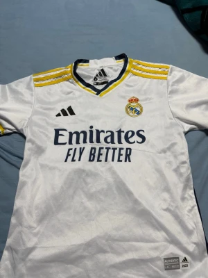 2 real Madrid t shirt  - Bilderna visar en vit Real Madrid matchtröja med gula och marinblå detaljer, Adidas-logga och Bellingham #5 på ryggen. Ingen sko finns med på bilderna.