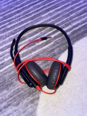 Kabelanslutna over-ear hörlurar med mikrofon - Svarta och röda kabelanslutna over-ear hörlurar med mikrofon och volymkontroll på sladden. Utrustade med 3,5 mm kontakt och USB-C adapter. Hörlurarna ser ut att vara i gott skick utan synligt slitage. Perfekt för samtal, gaming eller musik. Finns i Göteborg att hämta!