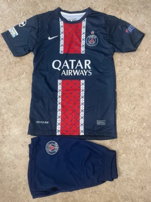 PSG fotbollströja och shorts set - Säljer ett Paris Saint-Germain fotbollskit från Nike med tröja och shorts. Tröjan är marinblå med röda och vita geometriska mönster, PSG-logga och Qatar Airways-tryck. Shorts är marinblå med PSG-emblem. Perfekt för fotbollsträning eller match.