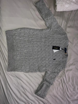 Grå kabelstickad tröja Polo Ralph Lauren - Kabelstickad grå tröja från Polo Ralph Lauren med klassisk rund hals och broderad logga på bröstet. Tröjan är långärmad och har ribbade muddar vid ärmslut och nederkant. Perfekt för dig som gillar stilren och tidlös design.