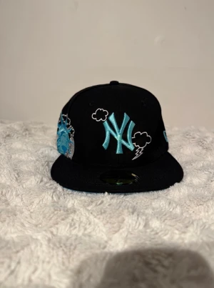 Svart New Era NY 59Fifty keps - Säljer en svart New Era 59Fifty keps med broderat NY-logga i ljusblått och vita moln och blixtdetaljer. På sidan finns frihetsgudinnan och stadssilhuett i blått och vitt. Baksidan har texten 'New York' broderad i ljusblått. Perfekt för dig som gillar streetwear. Lite mindre i storlek än vad det ska vara!