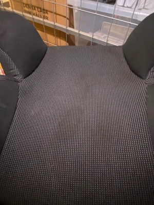 Svart rutig bilbarnstol med armstöd - Bilbarnstol i svart med rutigt mönster och vita sömmar. Stolen har höga sidor och armstöd, samt en bekväm sits i syntetmaterial. Passar perfekt för bilresor och har en modern, sportig look.