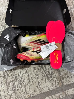 Adidas F50 fotbollsskor röd/gul - Säljer ett par Adidas F50 fotbollsskor med snygg design i rött, gult och svart. Skorna har snörning, platt sula med dobbar och är tillverkade i syntetmaterial. Medföljer röda innersulor och en matchande röd påse. Perfekt för dig som vill sticka ut på planen! Det finns en hål på toppen av skorna men inget annat. De går enkelt att fixa. Kan diskutera priset och de kommer med allt