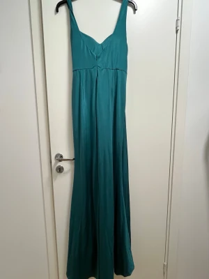 Turkos långklänning med drapering - Säljer en elegant turkos långklänning med breda axelband och v-ringad urringning. Klänningen har en snygg drapering framtill och är tillverkad i ett glansigt, mjukt material som ger ett lyxigt intryck. Perfekt för dig som vill sticka ut med en stilren och modern look.