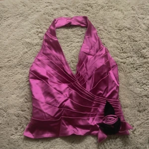 Rosa halterneck korsett med rosett - Säljer en glansig rosa korsett-topp i satin med halterneck och knäppning med tre knappar i nacken. Toppen har omlott-design, dekorativa veck och en svart rosett med blomdetalj framtill. Dragkedja i baksidan för enkel påklädning. Perfekt för att sticka ut.Sitter som en storlek S i midjan men större i bysten och runt nacken ungefär M