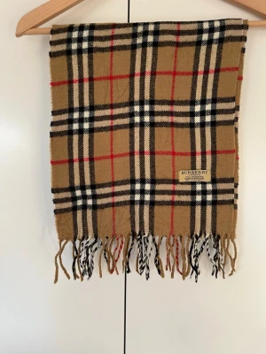 Burberry halsduk - Tidlös Burberry scarf i klassiskt rutmönster med fransar. Mjuk, varm och lätt att styla till både vardag och ytterplagg. | Material: 100 % kashmir | | Mått: ca 168 × 30 cm | | Mycket bra skick. |