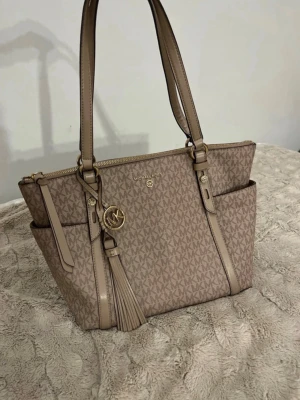 Beige handväska Michael Kors - Snygg beige handväska från Michael Kors med MK-logga över hela väskan. Väskan har två långa axelremmar, guldfärgade detaljer, tofs och dragkedja upptill. Praktiska sidofack och klassisk form, perfekt för dig som gillar stilrena accessoarer.