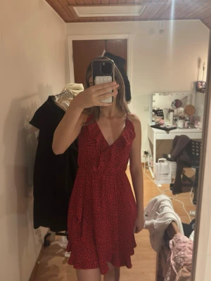 Röd leopardmönstrad omlottklänning - Säljer en röd kort omlottklänning med svart leopardmönster. Klänningen har volangdetaljer längs kanten, v-ringning och smala axelband. 