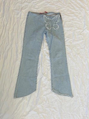 Ljusblå jeans med fjärilsdetalj - Unika ljusblå jeans med låg midja och bootcut-ben. Framtill finns en stor, fransad fjärilsdetalj som ger byxorna en cool och personlig vibe. Råa, fransiga kanter vid bensluten och dragkedja i sidan. Perfekt för dig som gillar statement-plagg och denim.