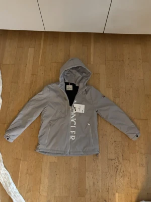 Grå vindjacka från Moncler med huva - Snygg grå vindjacka från Moncler med stor logga framtill, huva med snörning och två dragkedjefickor. Jackan har långärmad design och är tillverkad i polyester med ett mönstrat svart innerfoder. Perfekt för dig som gillar stilrena och sportiga plagg.