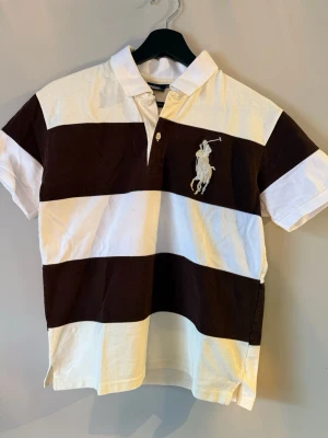 Randig polo piké från Ralph Lauren - Skick: 6/10. Storlek: M. Liten i storlek så skulle mer säga S. Ralph Lauren piké med breda bruna och vita ränder, stor broderad polospelare på bröstet och siffran 3 på ärmen. Pikén har krage och knappar framtill, tillverkad i mjuk bomull. Perfekt för dig som gillar old money stilen!