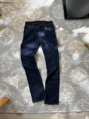 Nudie Jeans Thin Finn blå W29 L32 - Säljer ett par mörkblå Nudie Jeans Thin Finn med klassisk femficksdesign och snygga kontrastsömmar. Jeansen har en slim passform och är tillverkade av 98,5% ekologisk bomull och 1,5% elastan. Bakfickorna har Nudie Jeans signaturbroderi.