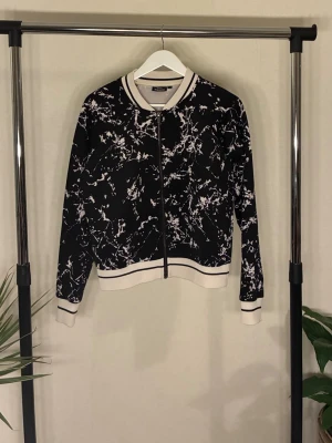 Bomberjacka  - Jackan har ett vitt abstrakt mönster, ribbade muddar och krage i vitt med svarta ränder samt dragkedja framtill.  