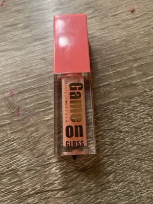 Game On Gloss från Makeup Mekka - Säljer ett läppglans från Makeup Mekka med namnet Game On Gloss. Glanset kommer i en genomskinlig fyrkantig tub med en korallrosa lock. Färgen ser ut att vara ljusrosa och ger en glansig finish. Perfekt för dig som vill ha snygga, glossiga läppar.