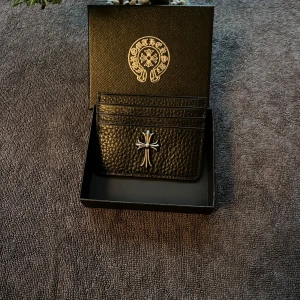 Svart plånbok från Chrome Hearts - Säljer en svart plånbok från Chrome Hearts i äkta skinn med grov textur. Plånboken har flera kortfack och är dekorerad med en silverfärgad korsdetalj på framsidan samt en metallskylt med loggan på baksidan. Kommer i original ask med logotyp.