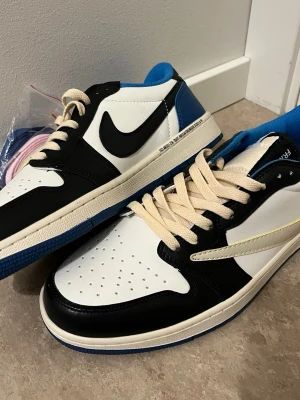 Nike Air Jordan 1 Low Travis Fragment - Nike Air Jordan 1 Low Travis Fragment med svart, vit och blå färg. Skorna har snörning, rund tå och platt sula. Materialet är skinn och detaljer som broderad logga på hälen och specialgrafik. Snygg kontrast mellan blå yttersula och vit/svart ovandel.