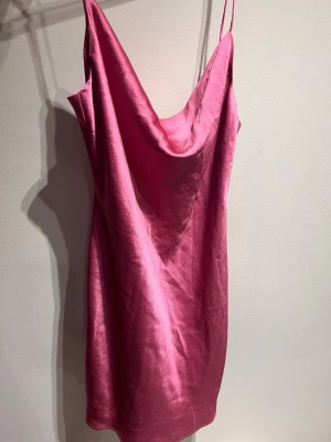 Rosa satin klänning med smala axelband - Säljer en snygg rosa klänning i satin med smala axelband och draperad halsringning. Klänningen är kort och har en mjuk, glansig yta. Aldrig använd 