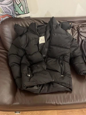 Svart Moncler pufferjacka - Säljer en svart pufferjacka från Moncler med ikonisk logga på ärmen och coolt serietidningsfoder. Jackan har hög krage, dragkedjor på fickorna och en ficka på ärmen. Perfekt för kalla dagar och riktigt snygg streetstil.