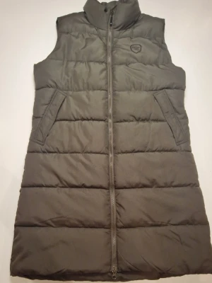 Lång väst i olivgrönt - Säljer en lång, quiltad väst från Tuxer i olivgrön färg. Västen har hög krage, två snedställda fickor och hel dragkedja framtill. 