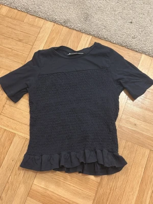 Mörkblå blus med volangkant - Säljer en mörkblå blus med korta ärmar och en smockad, rynkad framsida. Blusen har en rund halsringning och en söt volangkant längst ner💙storlek 146/152 men passar mig som brukar ha XS/S i storlek!