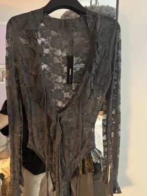 Grå spetsig bodysuit från Fashion Nova - En grå bodysuit från Fashion Nova med  spets och blommönster. Den har djup v-ringning, långa ärmar och snygga volangdetaljer längs kanten. Knytband framtill.  Oanvänd