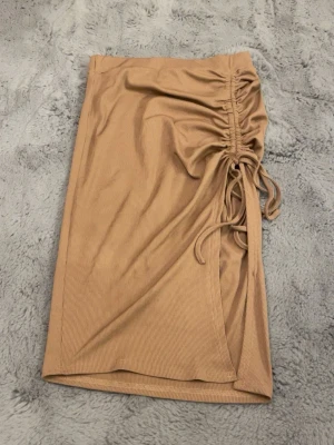 Beige ribbad kjol med snörning - Säljer en beige ribbad kjol med snygg dragsko så man kan göra slitsen längre och kortare och rynkdetalj på sidan. Kjolen har en omlottkänsla och är i mjukt, stretchigt material som sitter skönt. Perfekt för dig som gillar trendiga och stilrena plagg med lite edge. Kan eventuellt tänka mig gå ner i pris