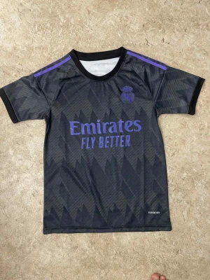 Real Madrid svart matchtröja - Säljer en svart Real Madrid matchtröja från Adidas med lila detaljer och geometriskt mönster. Tröjan har korta ärmar, rund hals och 'Emirates Fly Better' tryckt på bröstet. Perfekt för dig som älskar fotboll och vill ha en snygg supportertröja.