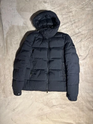  J.Lindeberg puffer jacka  - Svart pufferjacka från J.Lindeberg med huva och dragkedja framtill. Jackan har två sidofickor med dragkedja därav en är lite krånglig men går att fixa, den är tillverkad av polyamid och dun, vilket gör den riktigt varm. Perfekt för kalla dagar och har en clean, modern look. Hör av er vid fler frågor.