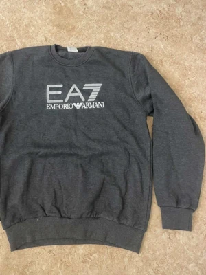 Mörkgrå sweatshirt från Emporio Armani - Snygg mörkgrå sweatshirt från Emporio Armani EA7 med stor vit logga framtill. Tröjan har rund hals, långa ärmar och ribbade muddar vid ärmslut och nederkant. Perfekt för en chill och stilren look.