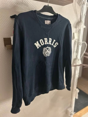 Mörkblå sweatshirt från Morris - Snygg mörkblå sweatshirt från Morris i storlek S med vit logotyp och emblem tryckt på bröstet. Tröjan har rund hals, ribbade muddar och är långärmad. Perfekt för dig som gillar klassisk och stilren design. Knappt använd och nypris runt 1600👌