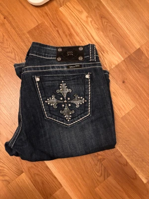 Miss Me mörkblå bootcut jeans med pärlor - Säljer ett par mörkblå jeans från Miss Me i bootcut-modell med snygga kontrastsömmar och pärlbroderad bakficka. Jeansen har låg midja, klassisk femficksdesign och detaljerade knappar. Perfekt för dig som gillar lite extra bling på dina jeans! Innerbenslängd 79cm