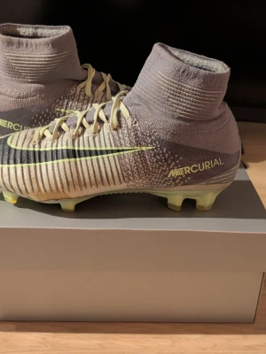 Nike Mercurial fotbollsskor beige - Säljer ett par Nike Mercurial fotbollsskor i beige och grått med svarta och gula detaljer. Skorna har snörning, ribbad ovandel och hög strumpkrage för extra stöd. Sulan är utrustad med dobbar för gräsplan. Materialet är syntet och stickad textil.