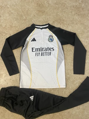 Ingen skor på bilderna - Bilderna visar ett Real Madrid träningsset från Adidas med långärmad tröja och svarta byxor, men inga skor finns med.