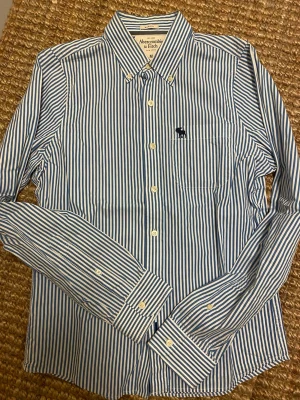Blåvit randig skjorta Abercrombie M - Snygg blåvit randig skjorta från Abercrombie & Fitch i storlek M. Klassisk krage, knappstängning och lång ärm. Bröstficka med broderad logga. Modellen är 'Muscle' och materialet känns som mjuk bomull. Perfekt för dig som gillar stilrena och fräscha skjortor.